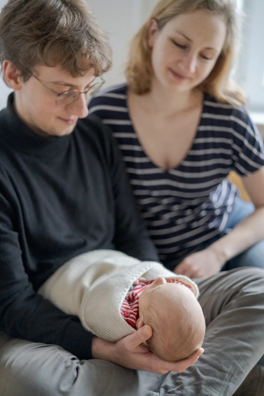 Mama und Papa sitzen bei einem Baby Shooting im Bett. Der Vater hält das Baby und die Mutter guckt es verliebt an.