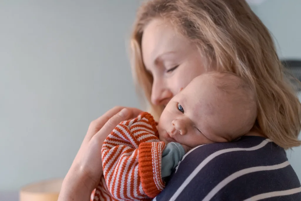 Eine Mutter trägt und kuschelt ihr Baby. Der Kopf des Babys liegt auf der Schulter der Mutter und die Mutter hat die Augen geschlossen.