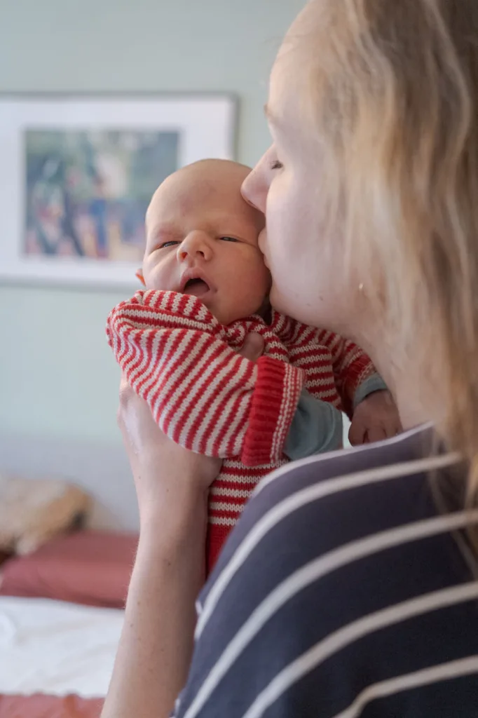 Die Mutter hält das Baby vor sich und gibt ihm einen Kuss, während sie im Schlafzimmer stehen und das Baby in die Kamera guckt.