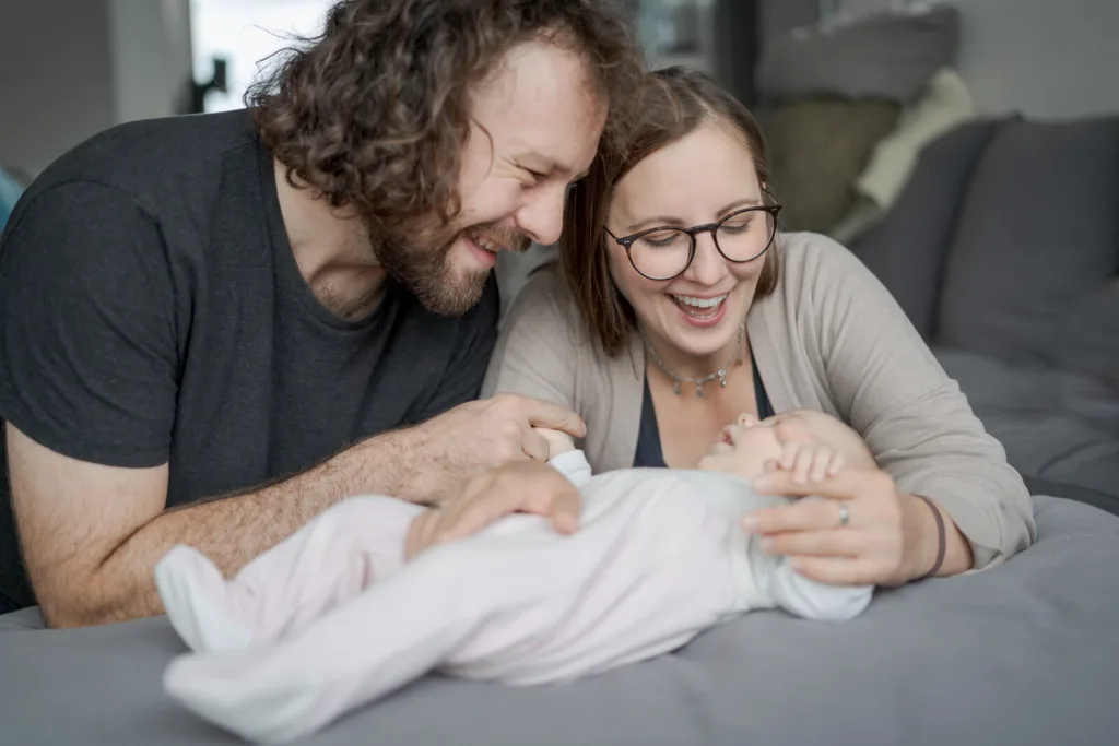 Ein Foto von einem Neugeborenenshooting. Es ist ein schwarz/weiß-Foto. Man sieht zwei junge Eltern, die Schulter an Schulter lehnen und sich ihr schlafendes Baby von oben angucken.