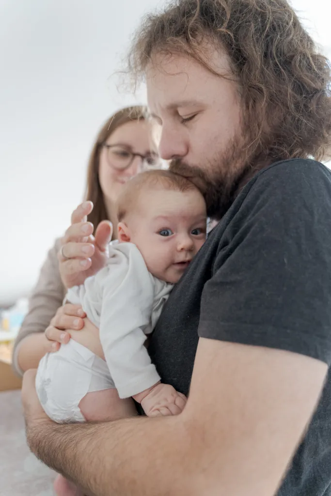 Ein Foto von einem Babyshooting. Zu sehen ist eine Familie mit Vater, Mutter und Baby, welches nur einen Body und eine Windel trägt. Der Vater hält das Baby, welches in die Kamera guckt, und gibt ihm einen Kuss auf den Kopf. Im Hintergrund sieht man die Mutter, die die beiden verliebt beobachtet.