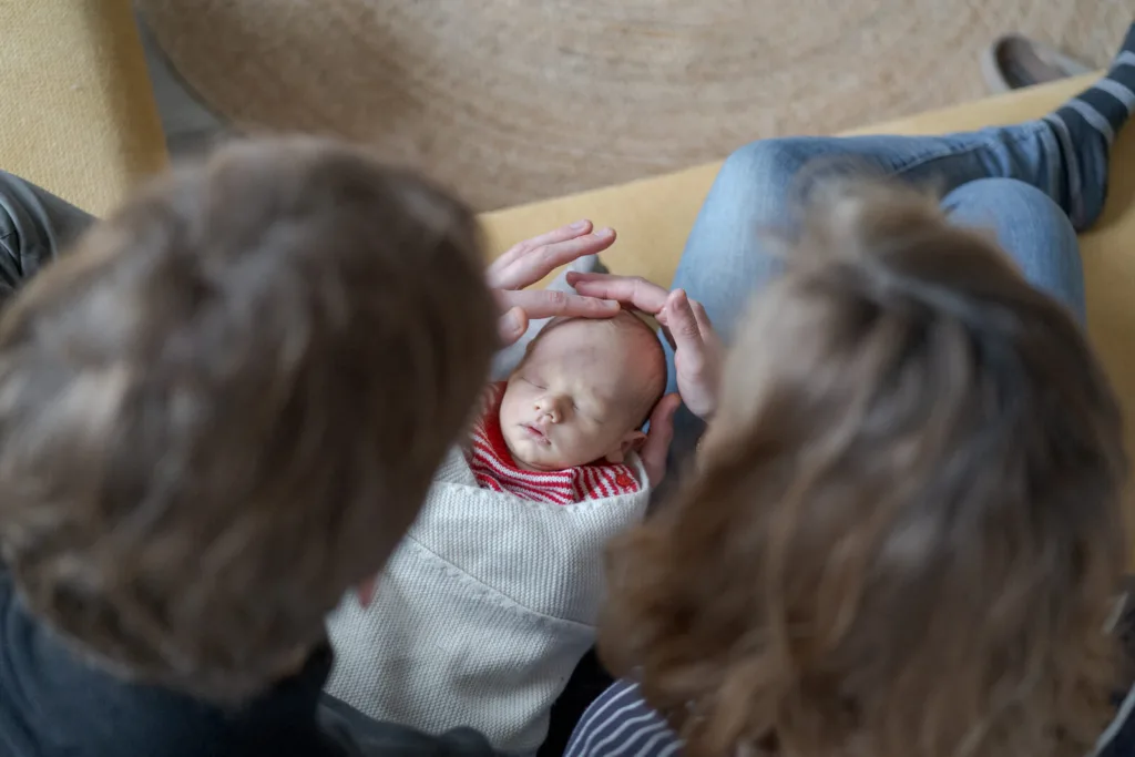 Das Foto wurde von oben gemacht. Die Eltern sitzen nebeneinander, halten das Baby im Schoß und streicheln es über den Kopf.