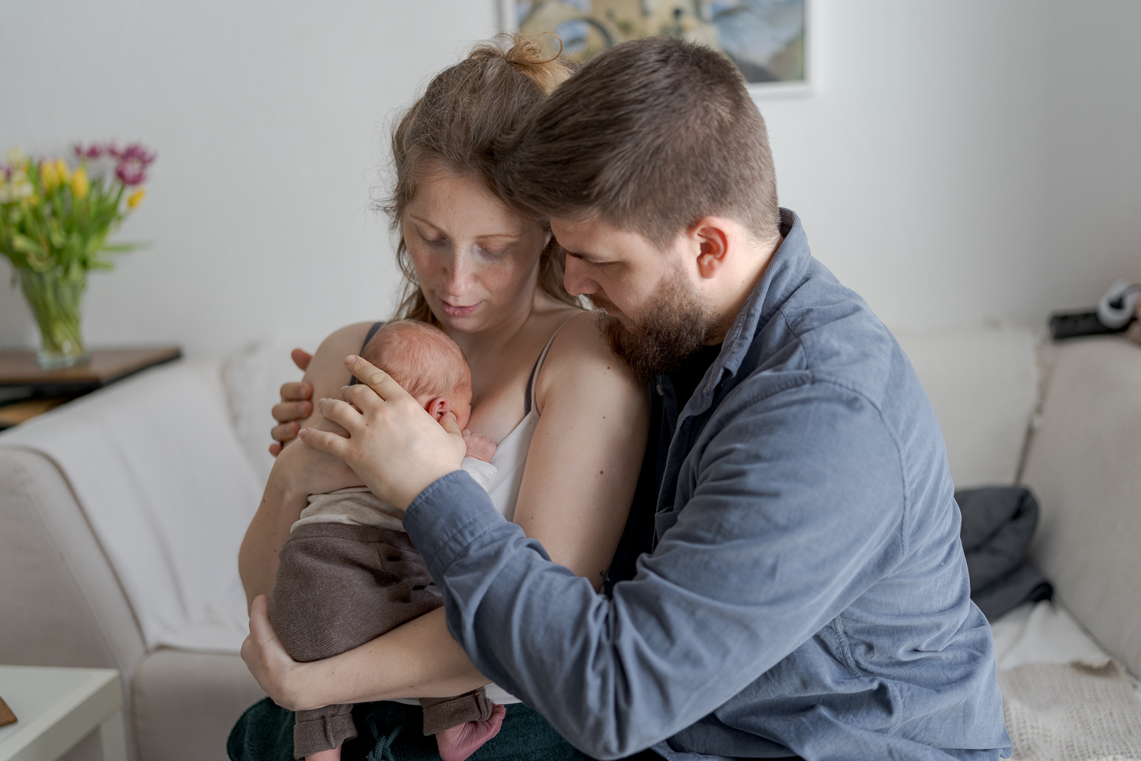 Es handelt sich um ein buntes Foto bei einem Babyfotoshooting. Es ist die ganze Familie zu sehen während sie auf der Couch sitzen. Die Mutter hält das Baby im Arm und der Vater umarmt beide gleichzeitig.