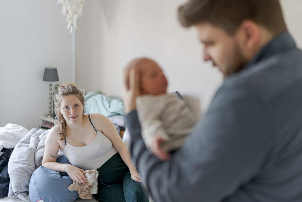 Es handelt sich um ein buntes Foto bei einem Babyfotoshooting. Die Mutter lehnt auf dem Bett und ist im Fokus. Der Vater hält das Baby und ist zusammen mit ihm verschwommen im Vordergrund zu sehen.