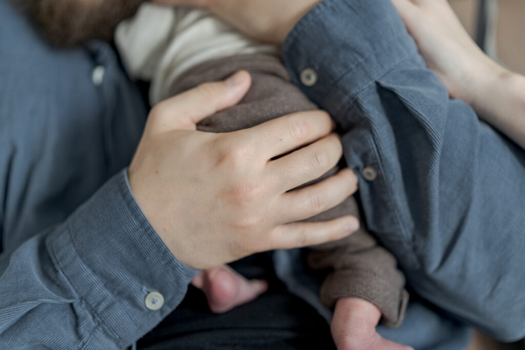 Es handelt sich um ein buntes Foto bei einem Babyfotoshooting. Es ist eine Detailaufnahme davon, wie der Papa sein Babyträgt und den Babypopo mit seiner Hand stützt.