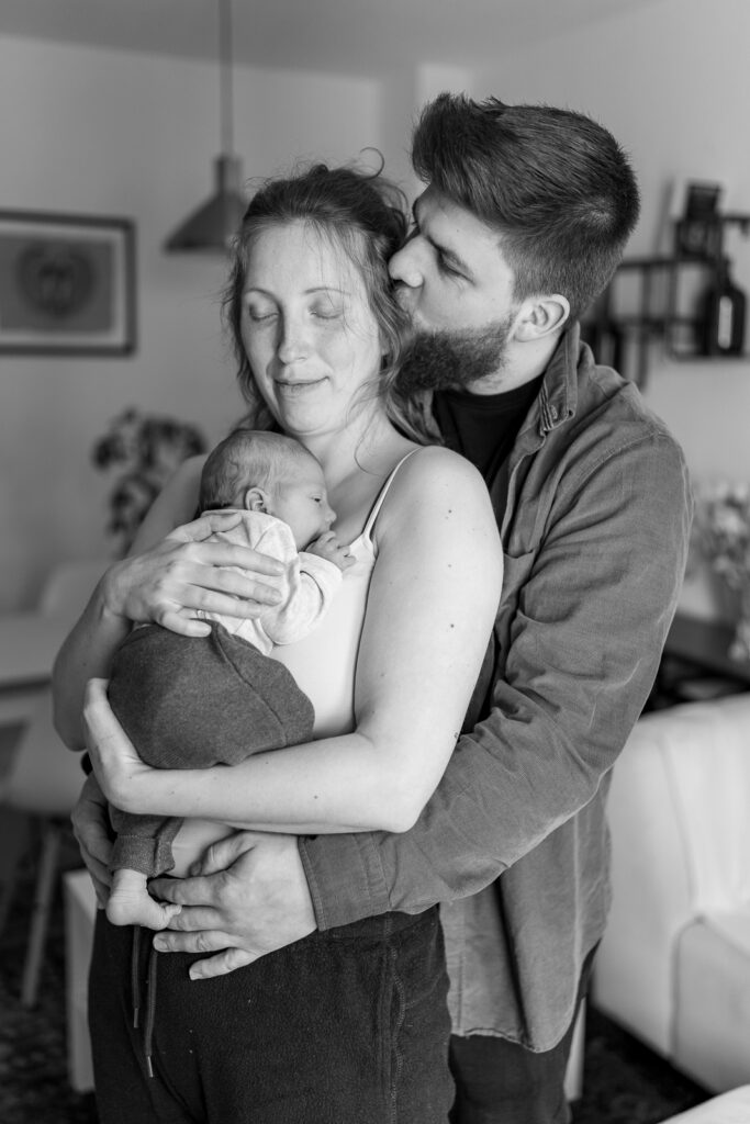 Es handelt sich um ein schwarzweiß Foto bei einem Babyfotoshooting. Es ist die ganze Familie zu sehen während sie zuhause im Wohnzimmer stehen. Die Mutter trägt das Baby und der Vater umarmt die Familie von hinten und gibt der Mutter einen Kuss während sie die Augen schließt.