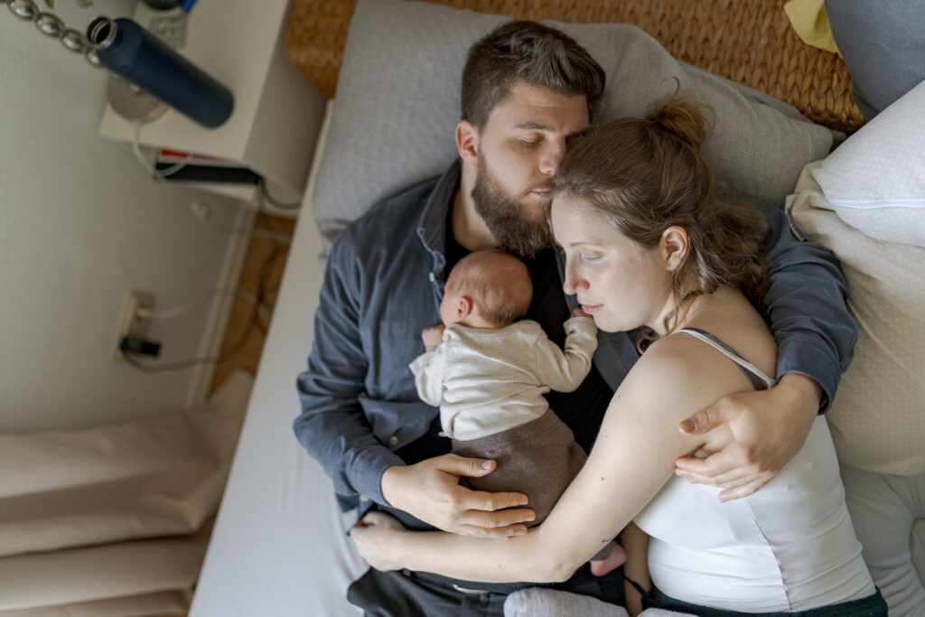 Es handelt sich um ein buntes Foto bei einem Babyfotoshooting. Die ganze Familie liegt im Bett und das Bild ist von oben aufgenommen. Das Baby liegt bäuchlings auf dem Bauch des Vaters und die Mutter kuschelt sich von der Seite ran.