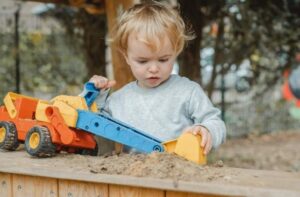 Ein Kleinkind spielt konzentriert mit einem Spielzeugbagger im Sandkasten.