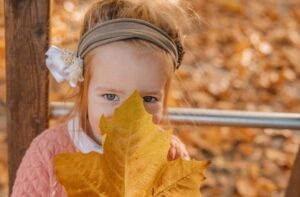Ein Mädchen hält ein großes Herbstblatt vor ihr Gesicht und schaut über den Rand hinweg in die Kamera.